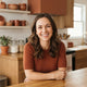 A friendly woman smiling in a bright, modern kitchen with warm terracotta and burnt sienna accents.